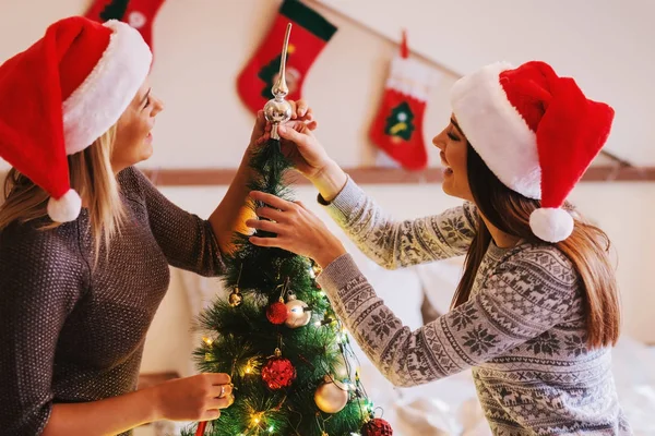两个头戴圣诞老人帽子的女性朋友装饰圣诞树 圣诞节假期概念 — 图库照片