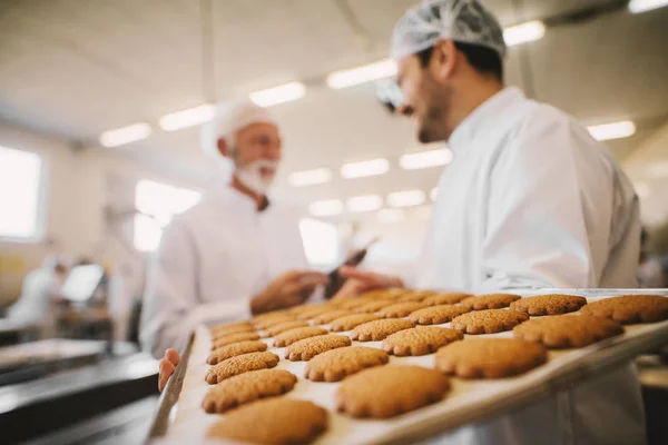 食品工場の大きな皿の新鮮なクッキーをクローズ アップ 無菌服バック グラウンドで話の つの男性従業員の画像をぼやけ — ストック写真