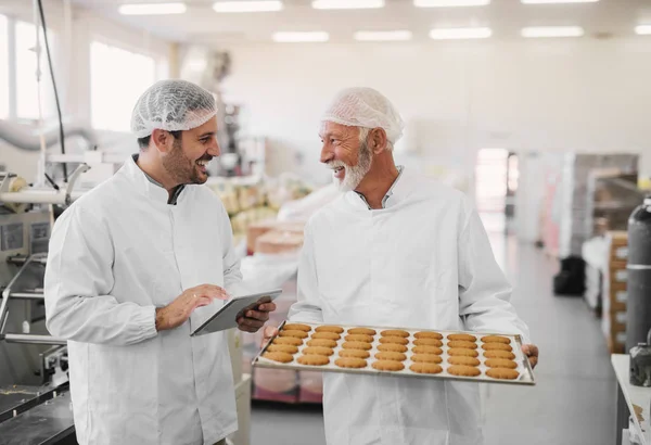 Picture of two employees in sterile clothes in food factory smiling and talking. Mature man is holding tray full of fresh cookies while the younger is holding tablet and checking production line.