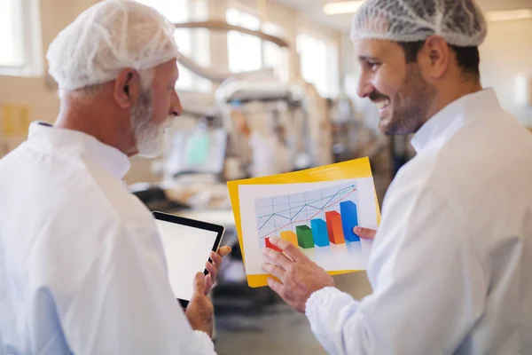 Two man standing and discussing. Younger man showing chart while senior watching and holding tablet. Food factory interior.