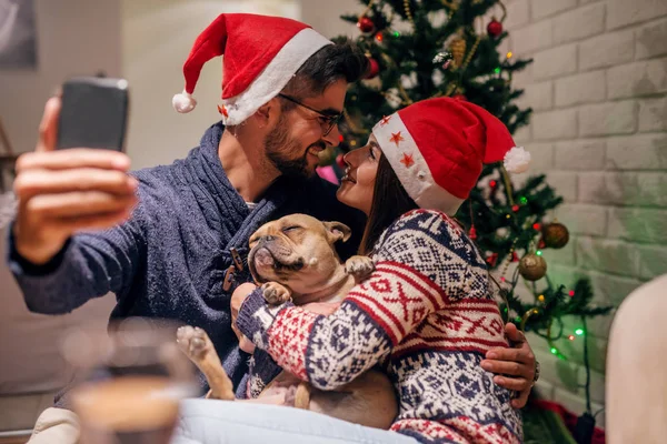 一对夫妇在圣诞树前和他们的狗一起进行自画像 戴在圣诞老人的帽子上 圣诞节假期概念 — 图库照片