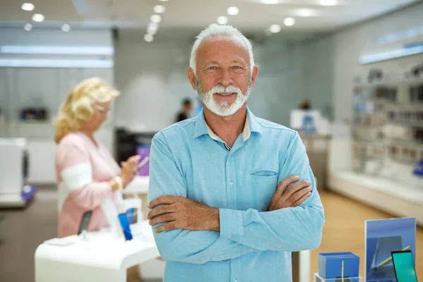 Bearded senior man standing in tech store with arms crossed and looking at camera.