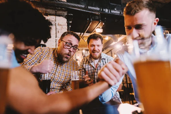 young happy bearded men drinking with beer in pub and having arm wrestling challenge