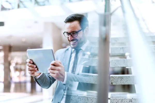 Smiling Businessman Using Tablet Reading Mail While Leaning Glass Lobby Stock Image