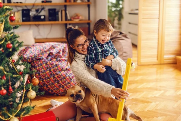 年轻的妈妈抱着狗在腿上玩的儿子 在他们旁边的圣诞树 家居内饰 圣诞节假期概念 — 图库照片