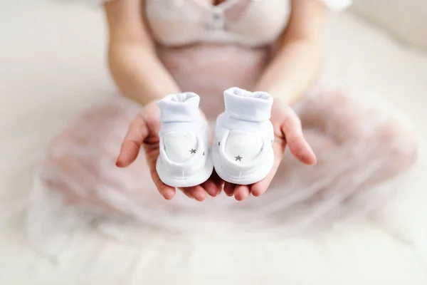 片方の手で赤ちゃんの靴を押しながら交差脚とベッドの上に座っている白人妊婦のクローズ アップ — ストック写真