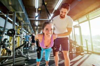 Determinate strong young personal trainer helping his female client to do TRX exercises correct. Client is looking happy and satisfied. clipart