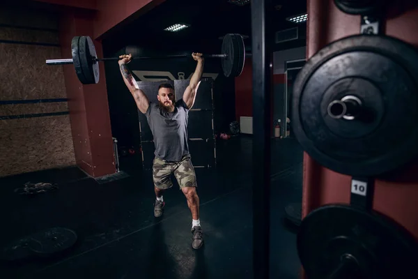 Muscular caucasian  bearded man holding barbell over his head doing barbell walking lunge exercise in crossfit gym. Weight plates in front.