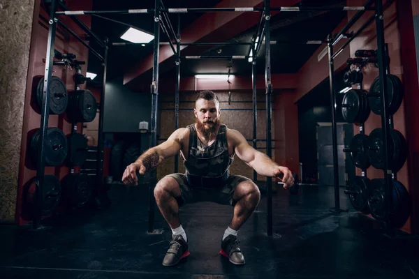 Muscular caucasian bearded man doing squats in military style weighted vest in crossfit gym.Cardio and strenght training. Weight plates and crossfit tires in background.