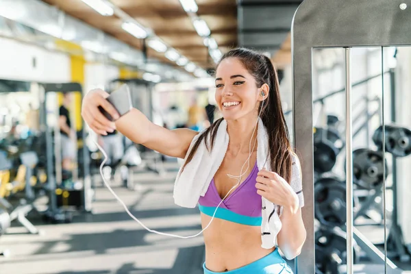 Caucasian woman with towel around neck and earphones in ears smiling and taking selfie while standing in a gym.