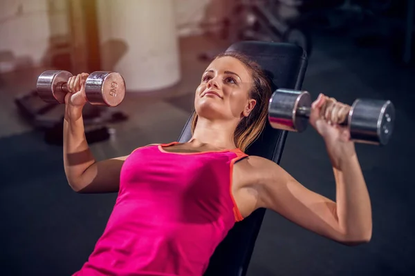 Young strong girl in a gym lying on bench and holding weighs in her hands.