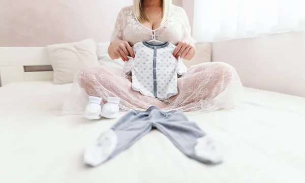 怀孕的白种妇女抱着婴儿的衣服坐在床上 双腿交叉在卧室 — 图库照片
