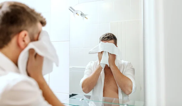 ボタンなしのシャツを着たハンサムなビジネスマンが 鏡の前に旅館の浴室に立ちながらタオルで顔を拭く 朝のルーチンコンセプト — ストック写真