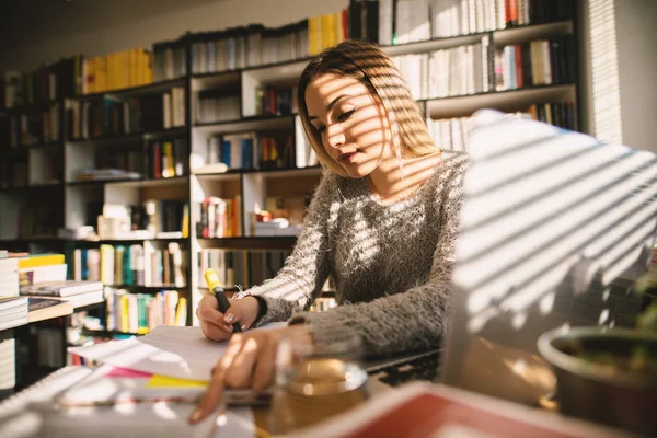 Cute young school girl sitting in library and taking notes. Sitting alone and studying in bright library.