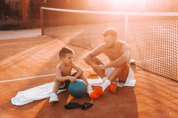 Shirtless father and son sitting on towels on court and resting from kicking ball. Summertime in the morning.