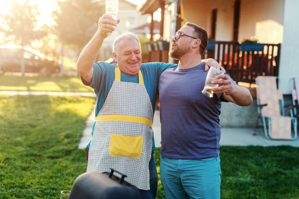 Cheerful father in law and son in law hugging and drinking beer while standing next to grill in backyard. Family gathering concept.