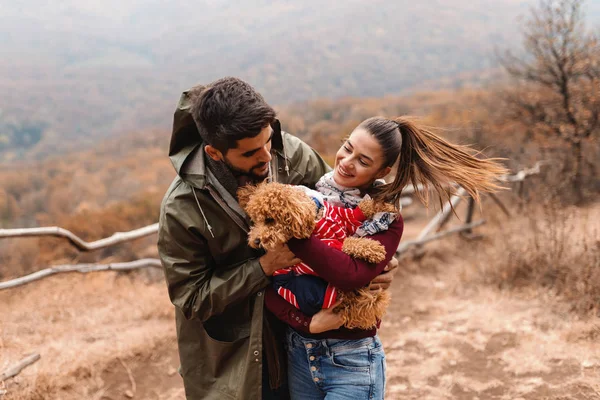 Beautiful woman holding dog while man cuddling him. In background forest and mountain. Autumn time.