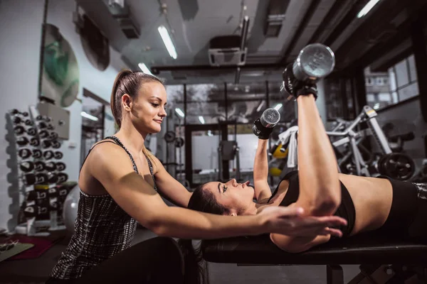Woman lifting dumbbells and lying down on bench while presonal treiner helping her.