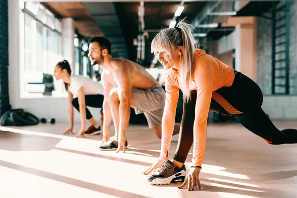 Small group of people with healthy habits doing stretching exercises on a gym floor. Selective focus on blonde woman. In background mirror.