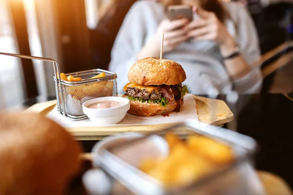 おいしいハンバーガー フライド ポテト 醤油皿の上の写真 スマート フォンを使用して背景の女性の — ストック写真