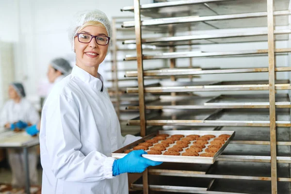 眼鏡とおいしいクッキー トレイをラックに入れて滅菌の制服を着た金髪の女性従業員の笑顔 — ストック写真