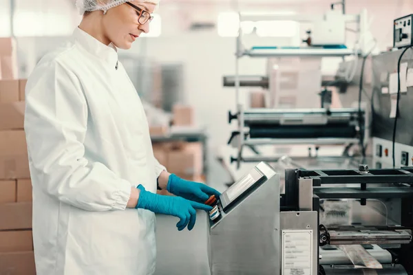 stock image Young female employee in sterile uniform and blue rubber gloves turning on packing machine while standing in food factory.