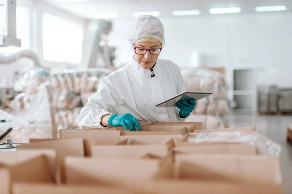 タブレットを保持しているとボックスの製品を数える滅菌の制服を着た若い白人の従業員 食品工場の内部 — ストック写真