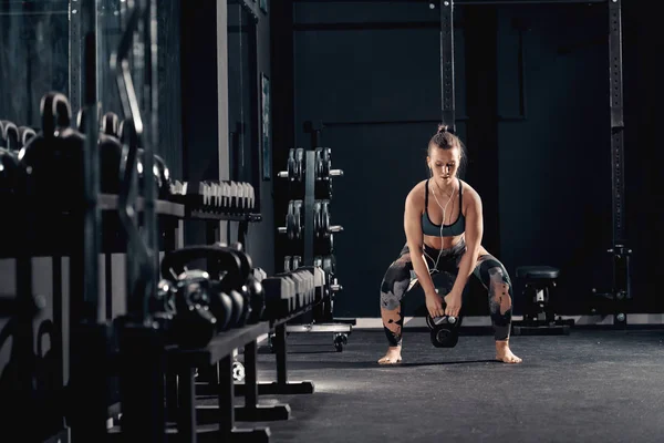 Caucasian muscular brunette with earphones in ears doing workouts with kettlebell in squat position. Night workout in gym concept.