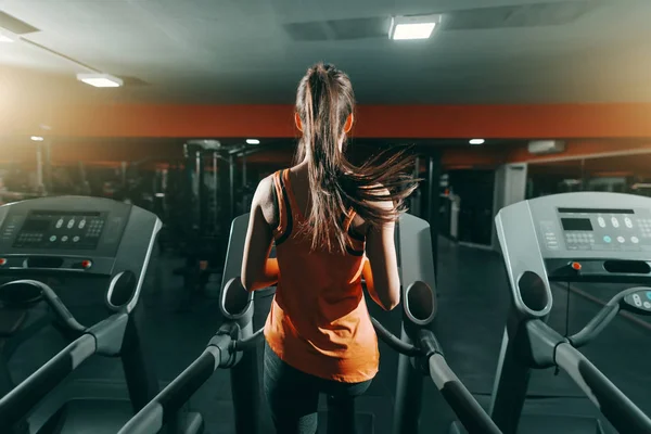 Slim brunette in orange sportswear and ponytail running on treadmill in gym at night. Backs turned. Be the girl who decided to go for it.