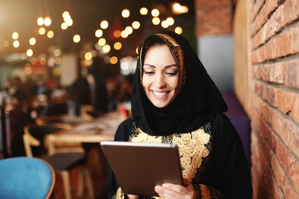 こぼれるような笑顔と美しいイスラム教徒女性が食堂に座りながらタブレットを使用して伝統的な摩耗に身を包んだ — ストック写真