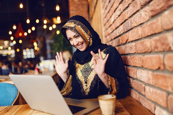 华丽的穆斯林妇女在传统的服装有视频通话在笔记本电脑上 而坐在自助餐厅和喝咖啡 — 图库照片