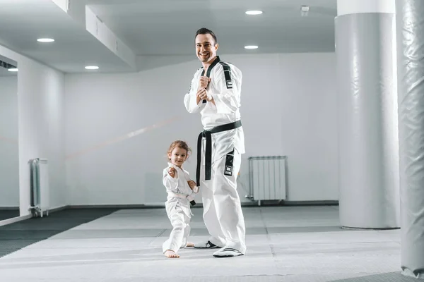 Hymyilevä Valkoihoinen Taekwondo Kouluttaja Poseeraa Pienen Tytön Kanssa Valkoisella Kuntosalilla — kuvapankkivalokuva
