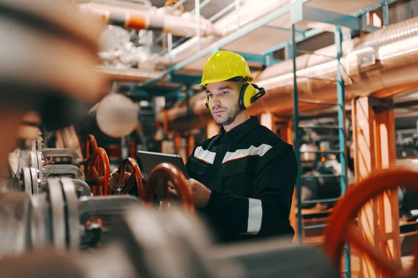 Young Caucasian hardworking heavy industry worker in protective suit and helmet using tablet and screwing valve. — Stock Image Young Caucasian Hardworking Heavy Industry Worker Protective Suit Helmet Using Stock Photo