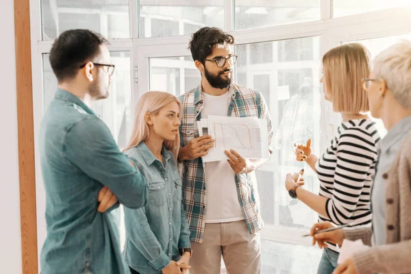 Business people standing next to window and discussing about project. Bearded man with eyeglasses holding business plan. Start up business concept.