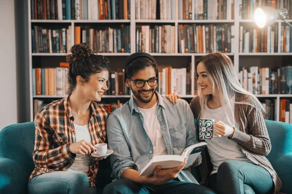 多文化学生図書館と本の話でソファーに座っていた 女性がコーヒーを飲みながら本を抱きかかえた 背景の本棚で — ストック写真