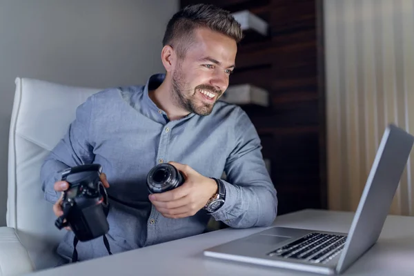 ハンサムな笑顔白人ひげを生やした手でカメラを保持して ノート パソコンを見て 夜遅くまでオフィスに座っている青いシャツを着たカメラマン — ストック写真