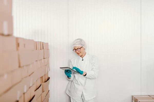金发碧眼的女性员工穿着无菌制服 在食品厂的箱子旁边站着用片剂进行后勤 — 图库照片