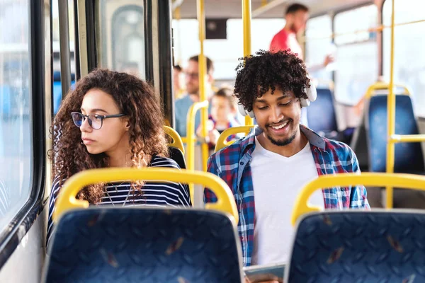 两个年轻人坐在城市公共汽车上 女孩透过窗户看 而男人有耳机的耳朵和使用平板电脑 — 图库照片