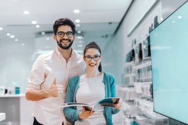 Young attractive multicultural couple looking for new plasma tv. Woman holding brochure while man holding thumbs up. clipart