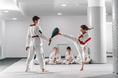 Caucasain boy and girl in doboks having taekwondo training at gym. Girl kicking while boy holding kick target. In background their friend sitting with legs crossed and watching them. clipart