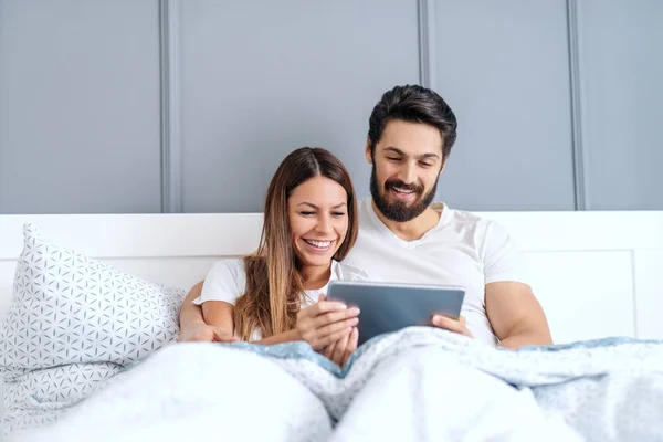 パジャマで幸せな白人のカップルと歯の笑顔でベッドに横たわって タブレット上のビデオを見て ベッドルームインテリア — ストック写真