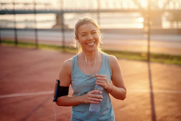 Smiling Caucasian pretty blonde sporty woman standing on court whit phone case around arm, earphones in ears and water in hands. A champion is afraid of losing. Everyone else is afraid of winning.