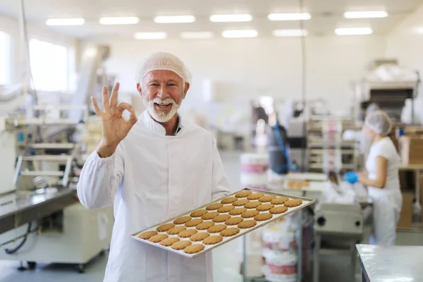 Funcionário Sênior Uniforme Estéril Segurando Caçarola Com Biscoitos Mostrando Sinal — Fotografia de Stock