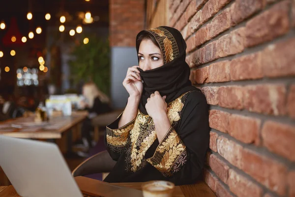 华丽的穆斯林妇女覆盖她的脸与围巾 而坐在咖啡店 桌子上有笔记本电脑和咖啡 — 图库照片