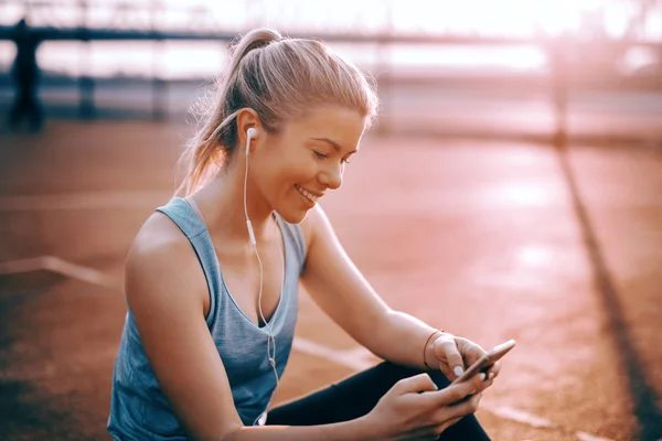 Cute smiling Caucasian blonde sporty woman sitting on the court with earphones in ears and using smart phone.