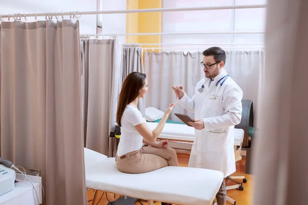 眼鏡と青いユニフォームを着た深刻な白人医師は タブレット上で彼の女性患者治療計画に示しています 病院の内部 — ストック写真