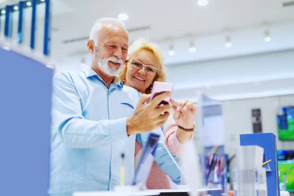 ハイテク店に立って 新しいスマートフォンを選択するかわいい笑顔のシニアカップル — ストック写真