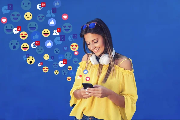 青い背景の前に立っている間 ソーシャルメディアのためにスマートフォンを使用して 首の周りにサングラスとイヤホンを持つ黄色のブラウスで笑顔かわいい十代の女の子 — ストック写真