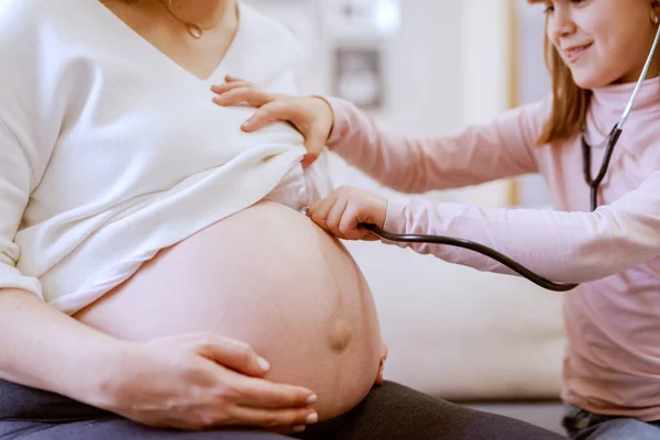 美しい白人の妊娠中の母親は 彼女の娘とリビングルームに座っています 聴診器の赤ちゃんの心拍で聞く娘 リビングルームのインテリア — ストック写真