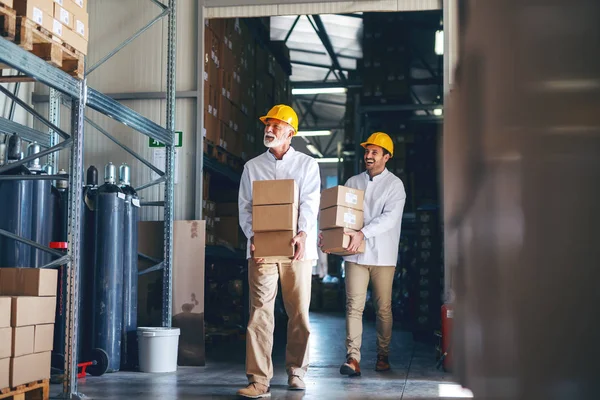 两名蓝领工人身着白色制服 头上戴着黄色头盔 在仓库里安置重箱子 — 图库照片